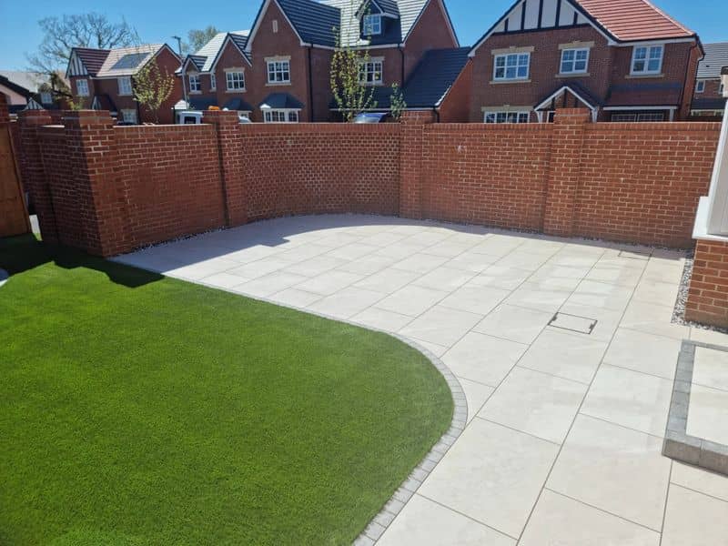 Rear garden with curved artificial grass and light porcelain patio against red brick wall
