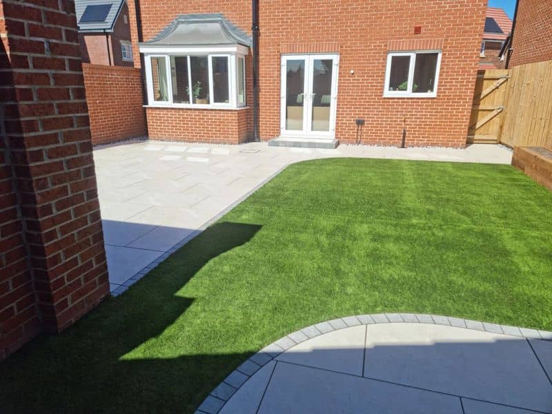 Finished rear garden with bright artificial grass and light porcelain patio by red brick house