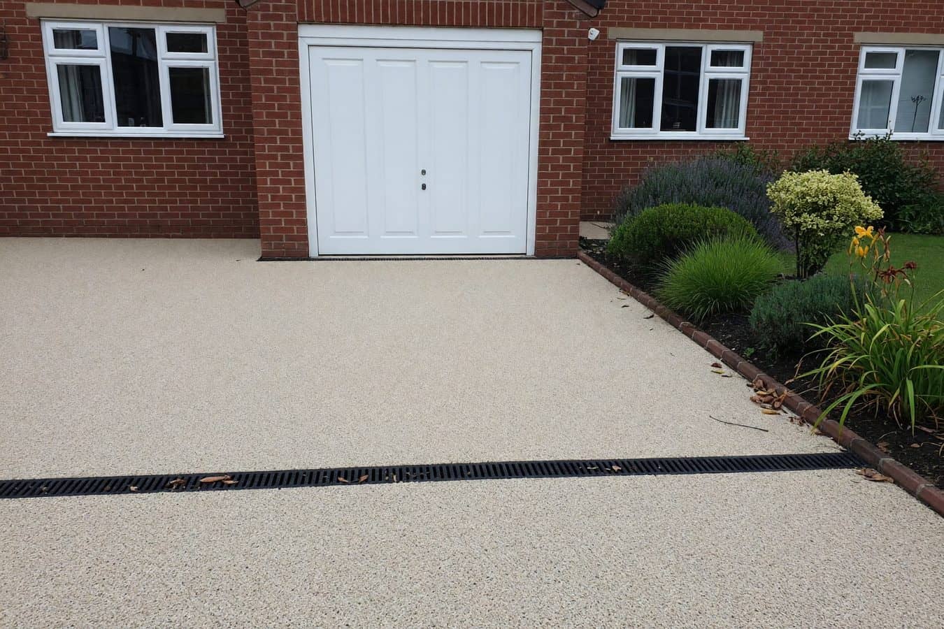 Channel drain set flush in a pale resin bound driveway at a white PVCu garage door on a Lancashire brick house